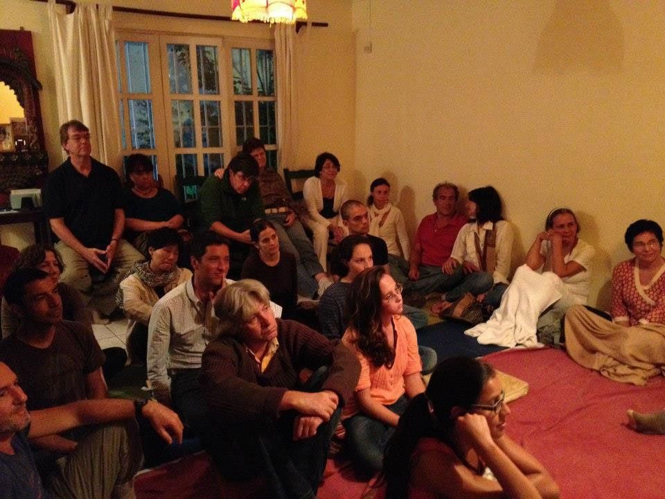 People sitting on a red floor, listening attentively. They are gathered in a well-lit room with windows.