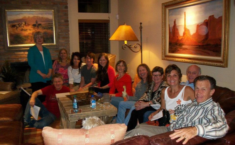 Group of people seated and standing in a living room, smiling, with landscape paintings on the wall.