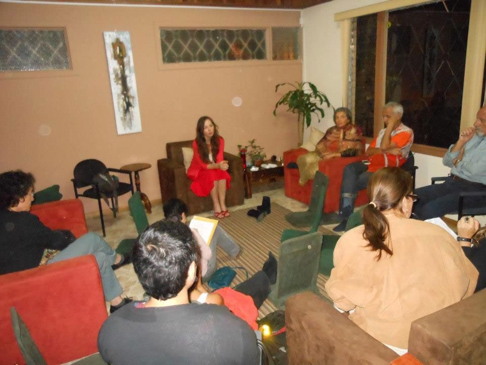 People seated in a living room, listening to a woman in a red dress. Interior lit, neutral tones.
