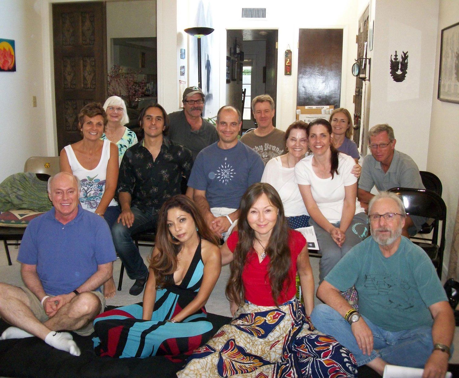 Group of people indoors, some sitting, others standing. Smiles, various clothing, neutral expressions, in a bright room.