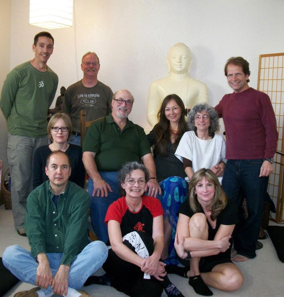 Group photo: people in a room, Buddha statue in background. Some sit, some stand, all smiling.