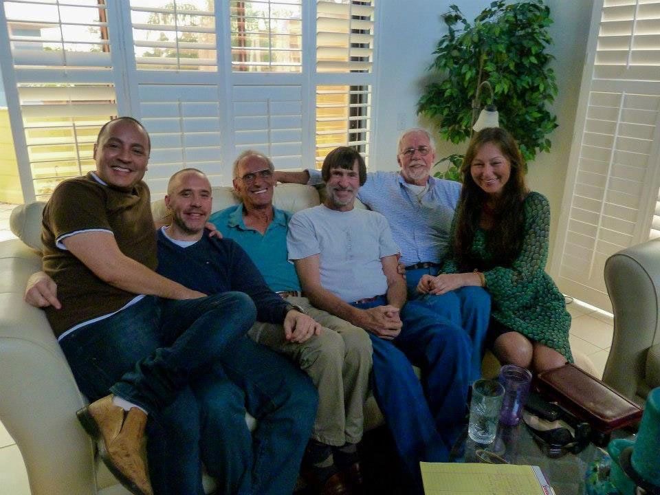 Group of six people smiling, seated together on a cream-colored couch, in a room with shutters, a plant, and a coffee table.