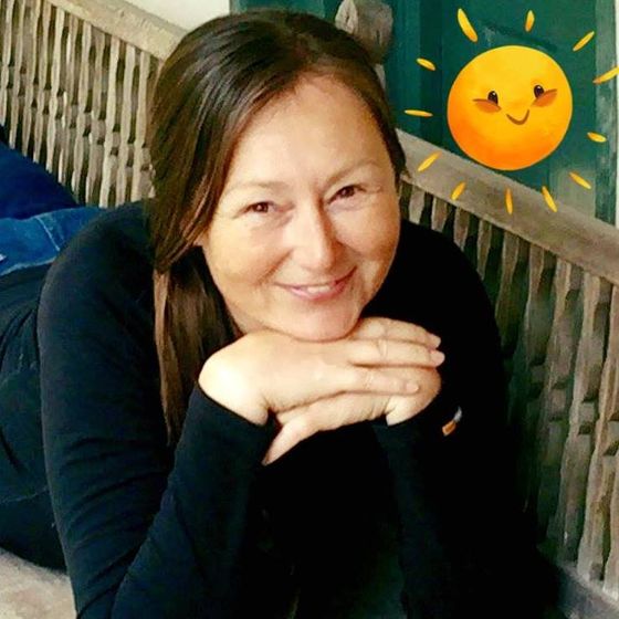 Woman smiling, leaning on hands, on a wooden bench, with a cartoon sun graphic.
