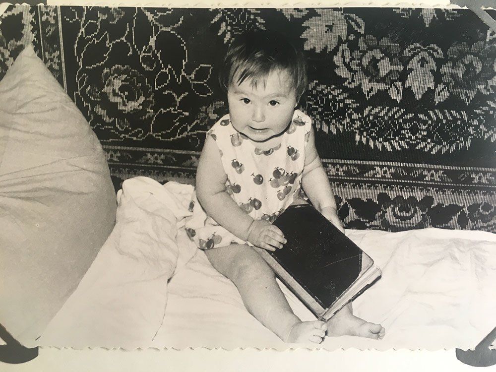 Baby sitting on a bed, holding a book. Floral patterned cloth behind the baby.