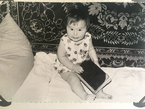 Baby sitting on a bed, holding a book. Floral patterned cloth behind the baby.