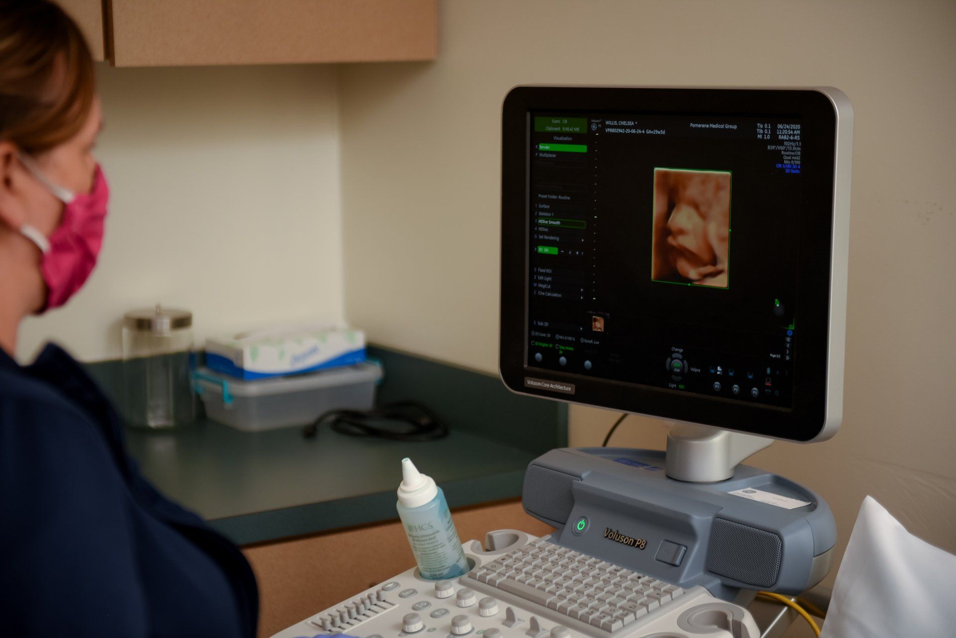 A healthcare worker wearing a pink mask examines an ultrasound image of a baby on a screen.