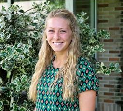 Smiling blonde woman in a green and black patterned shirt poses outside in front of a green bush.