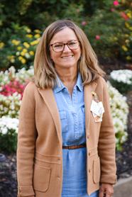 Woman with glasses smiles while wearing a brown jacket over a denim dress, standing in front of colorful flowers.