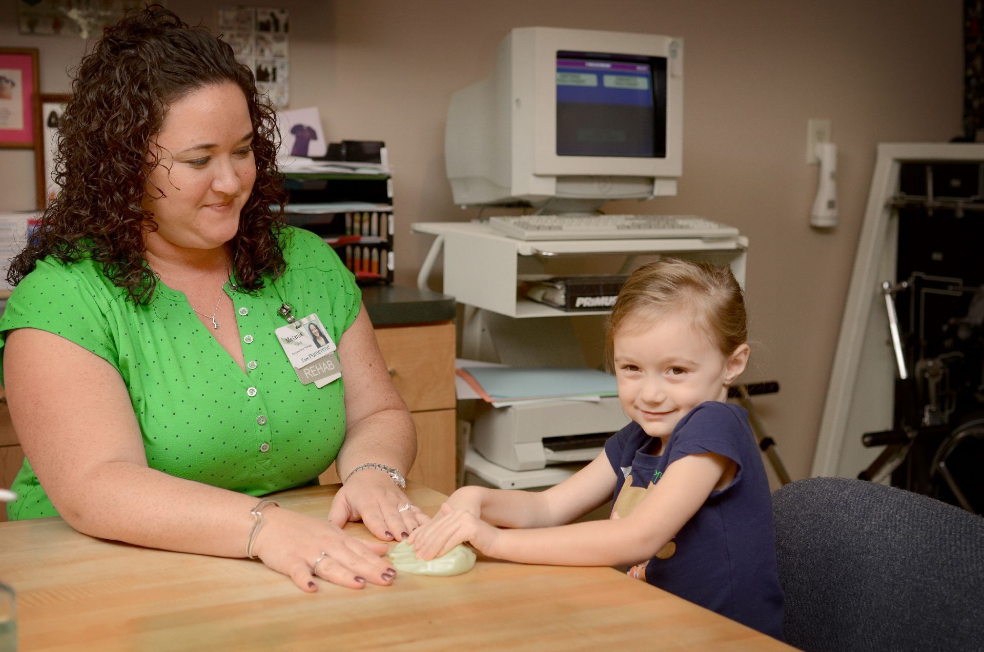 Woman in green shirt and young girl smiling, playing with clay on a wooden table in a room with a computer and printer.