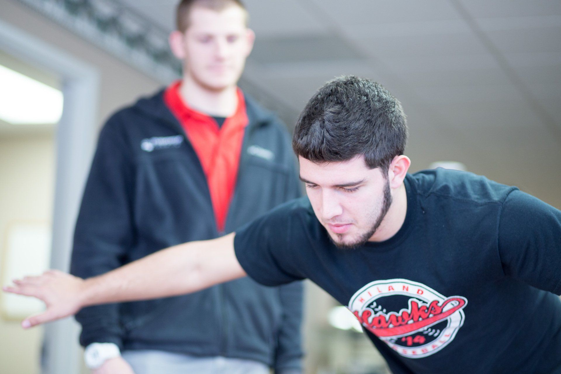 A man in a black t-shirt balances with one arm extended while another man watches. They're indoors, likely a gym.