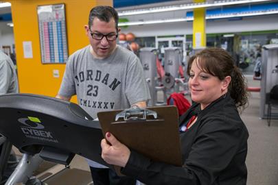 A fitness instructor assists a man on a treadmill, taking notes on a clipboard in a gym setting.