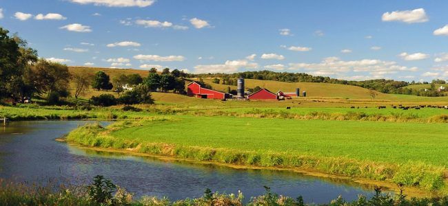 Scenic farmland with a red barn and silo under a blue sky. A river flows through a green field, with trees on the horizon.