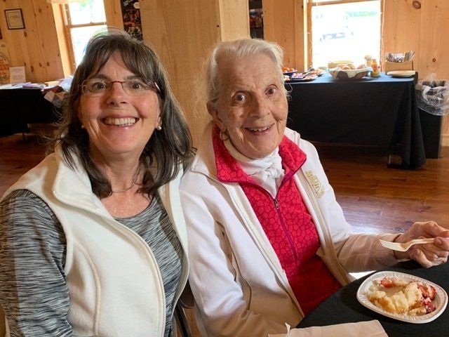 Two women smiling, seated at a table. One wears a white vest, the other a white jacket.
