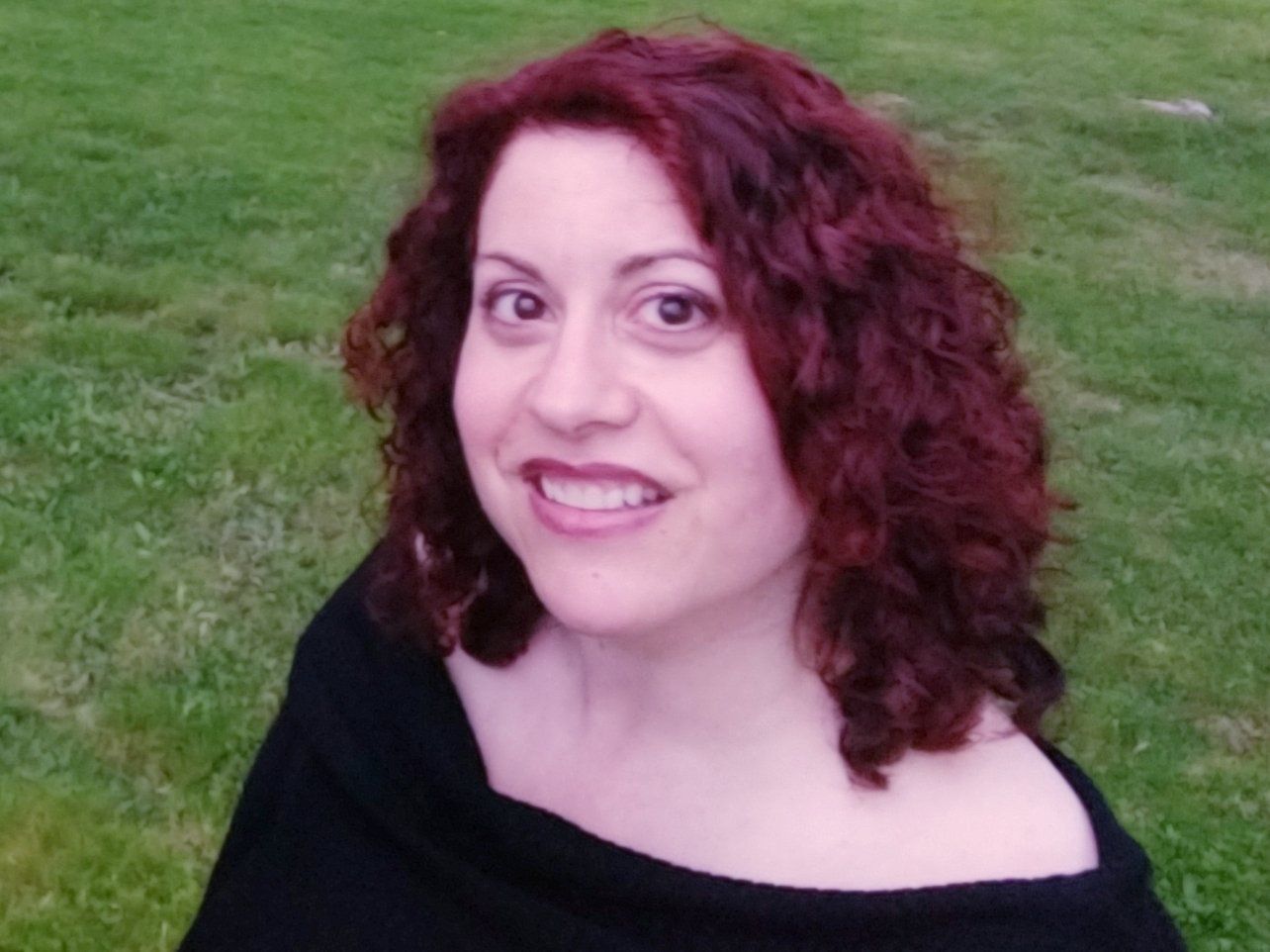 Woman with red curly hair smiles against a green grassy background. She wears a black top.