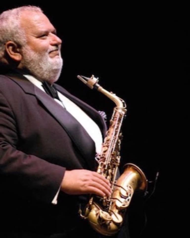 Saxophonist in a tuxedo, playing the instrument. Black background, smiling.