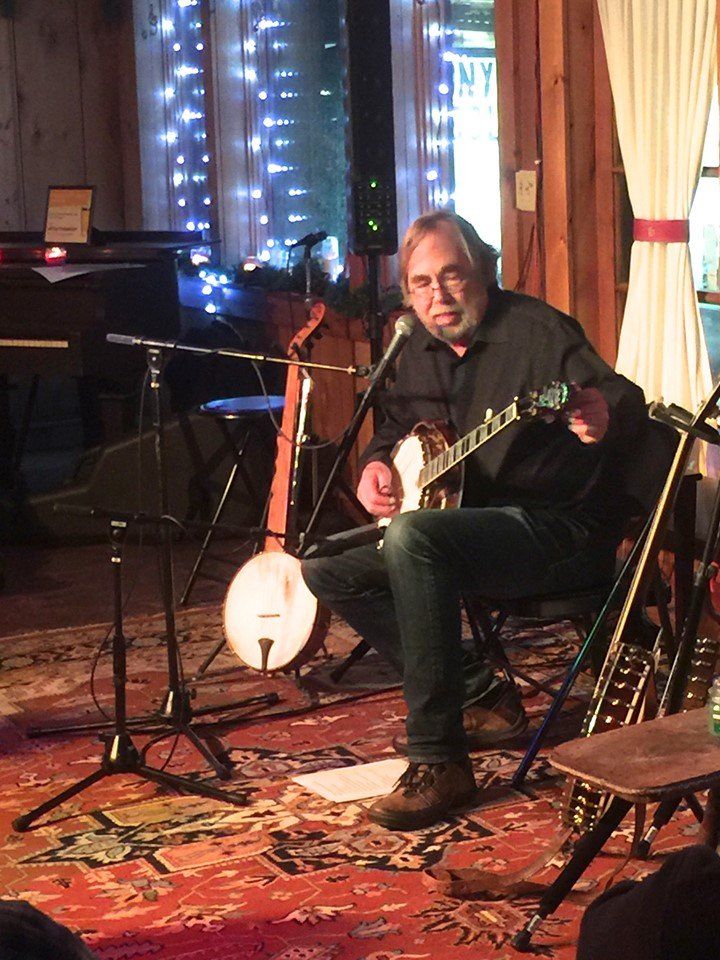 Man playing banjo, seated on stage with microphones, instruments, and fairy lights.