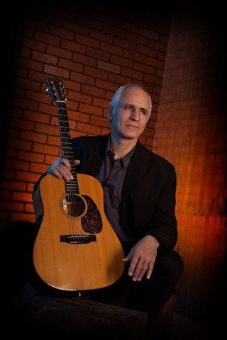 Man in black suit holds acoustic guitar, poses in front of a brick wall.