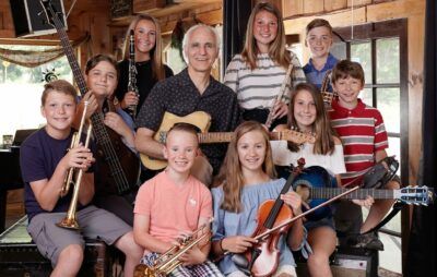 A group of musicians, adults, and children, with instruments indoors: guitars, trumpets, violin, and clarinet.