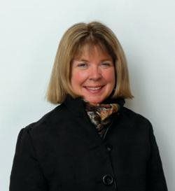 Woman with blonde hair smiling, wearing a black coat and patterned scarf against a white wall.