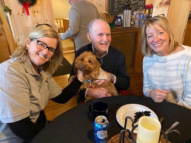 Three people and a dog seated at a table. The man holds the dog, a woman pets it, and another woman smiles. Indoor setting.