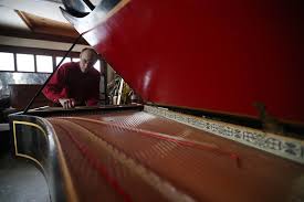 Man working on the inside of a grand piano. Red instrument top open, strings visible. Garage setting.