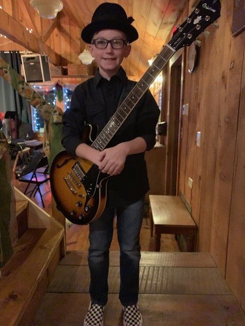 Young person in a black hat, shirt, and jeans holding a guitar. Inside a wooden-walled room.