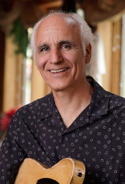 Man smiling, holding guitar, wearing patterned shirt. Light background, soft natural lighting.