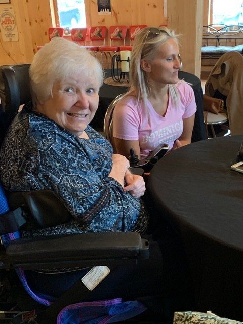 Woman in wheelchair smiles, beside another woman at a table. Wooden interior.