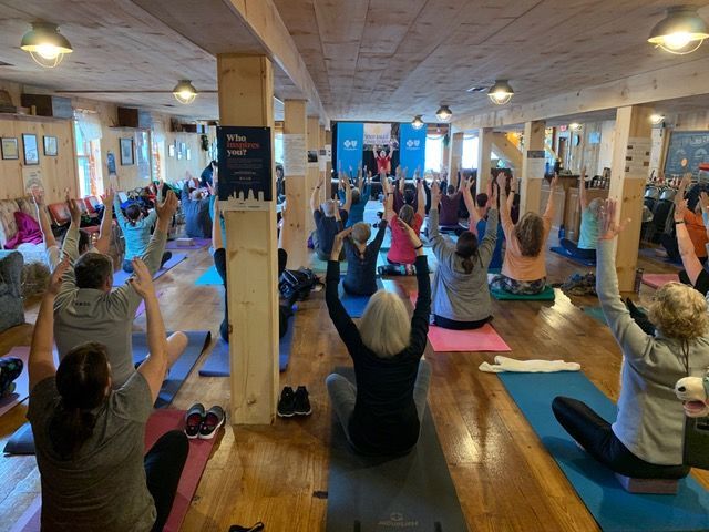 People in a large room doing yoga, arms raised. Bright, wood-floored space.