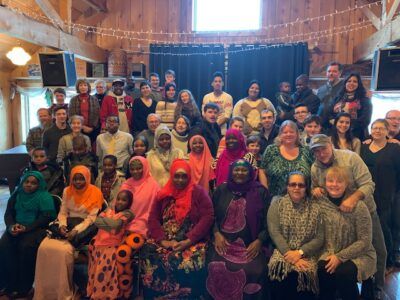 Large group of people indoors, smiling and posing. Some wear head coverings. Wooden interior with stage.