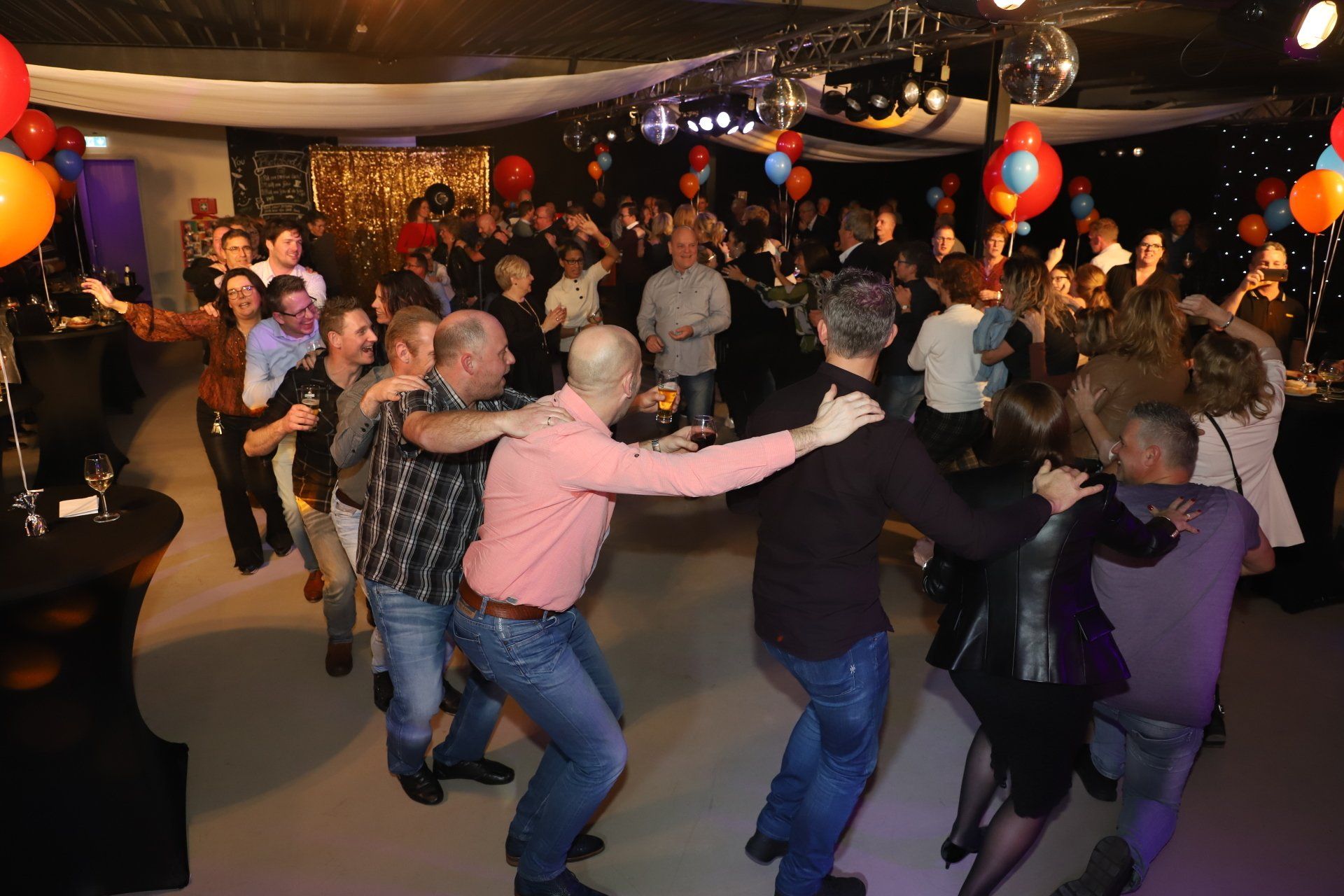 Mensen dansen op een feest, sommigen in een rij, met ballonnen en discoballen.