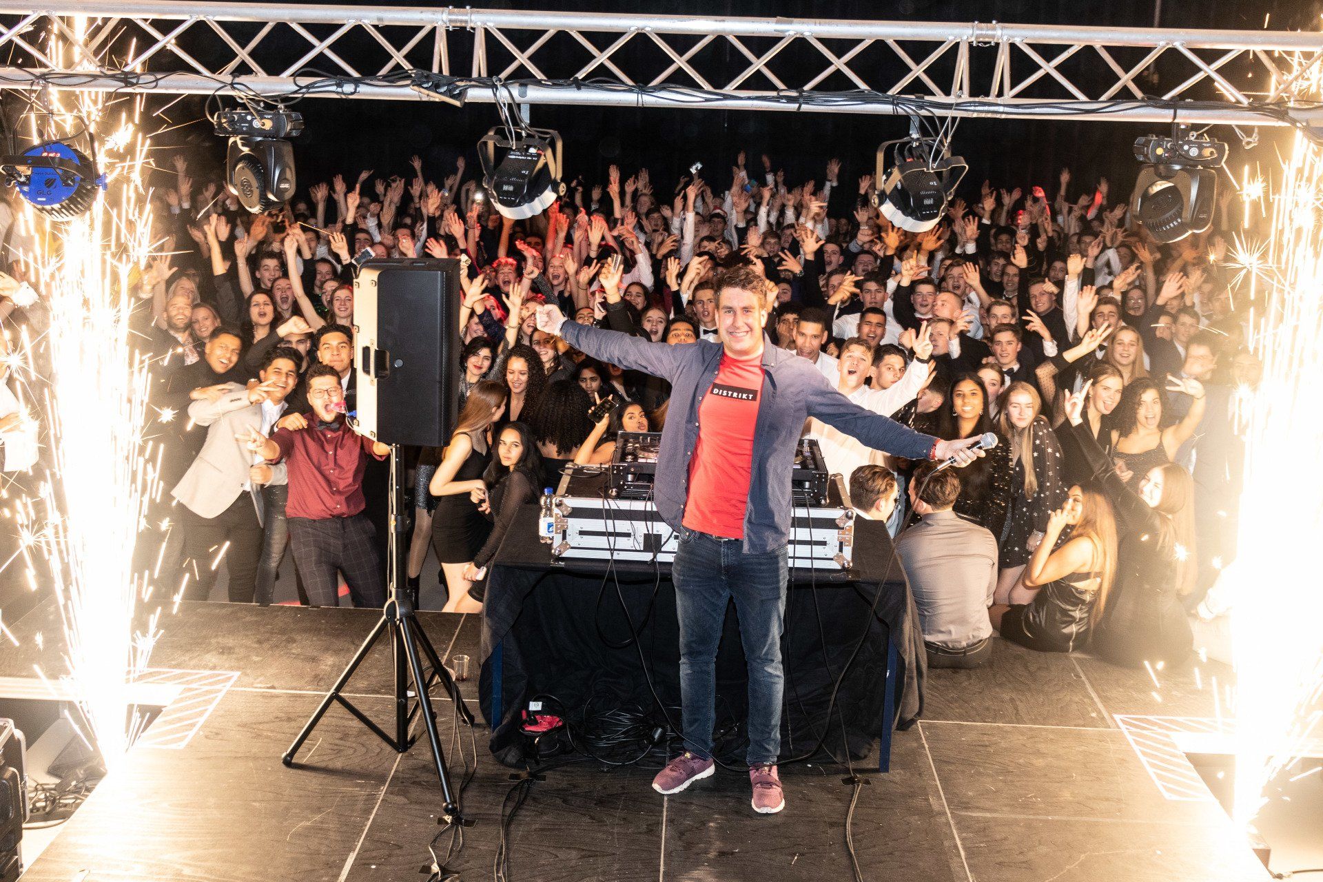 Een dj staat met uitgestrekte armen op het podium, omringd door een juichende menigte tijdens een avondevenement, met vonken op de achtergrond en podium effecten.