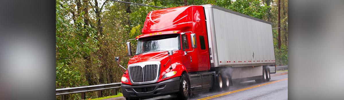 Red semi-truck driving on a wet road, surrounded by green trees, and a silver trailer.