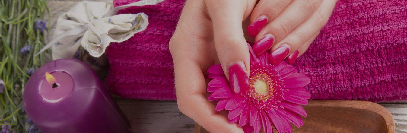 Mani con unghie rosa che tengono un fiore rosa, accanto a una candela viola e un asciugamano.
