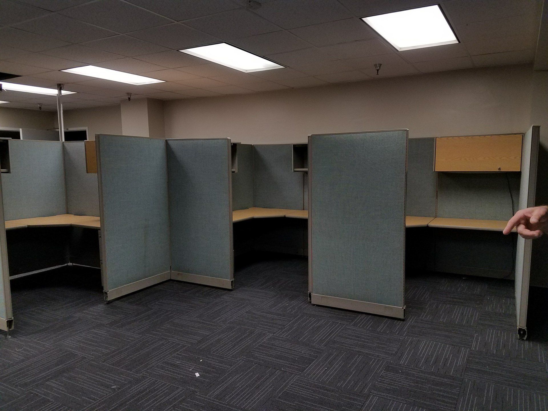 Empty office cubicles with gray dividers and desks, under fluorescent lights.
