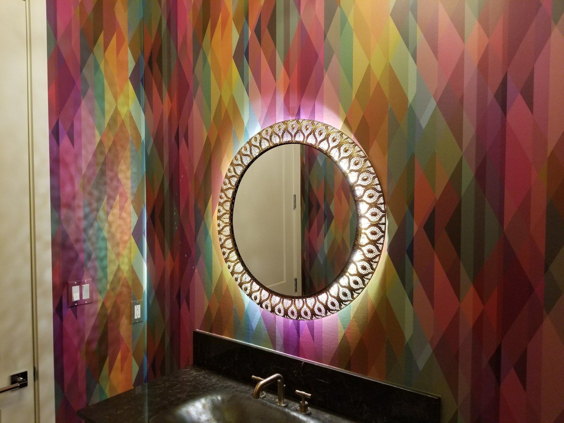 Bathroom with colorful geometric wallpaper, a round mirror, and a black countertop.