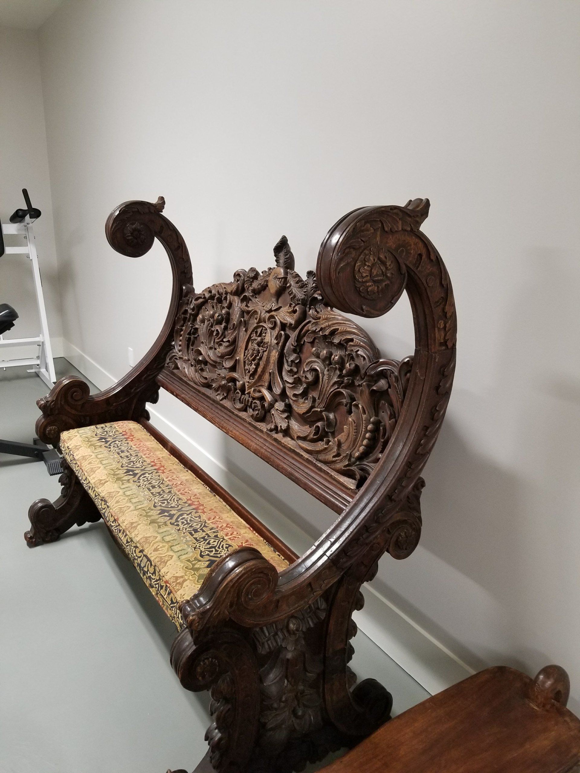 Ornate wooden bench with carved back and scroll arms, padded seat against a neutral wall.