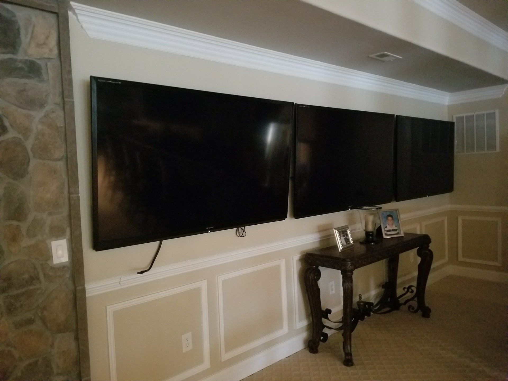Three mounted TVs on a wall above a table in a room with wainscoting.