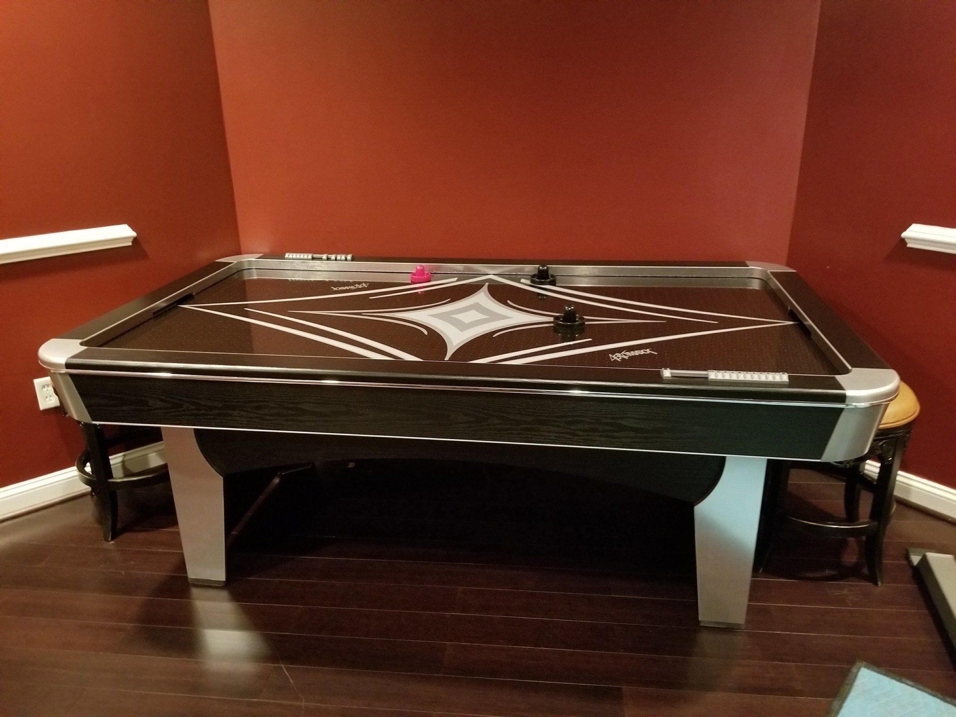 Air hockey table in a corner, with a red playing surface, black and silver trim, and grey legs.