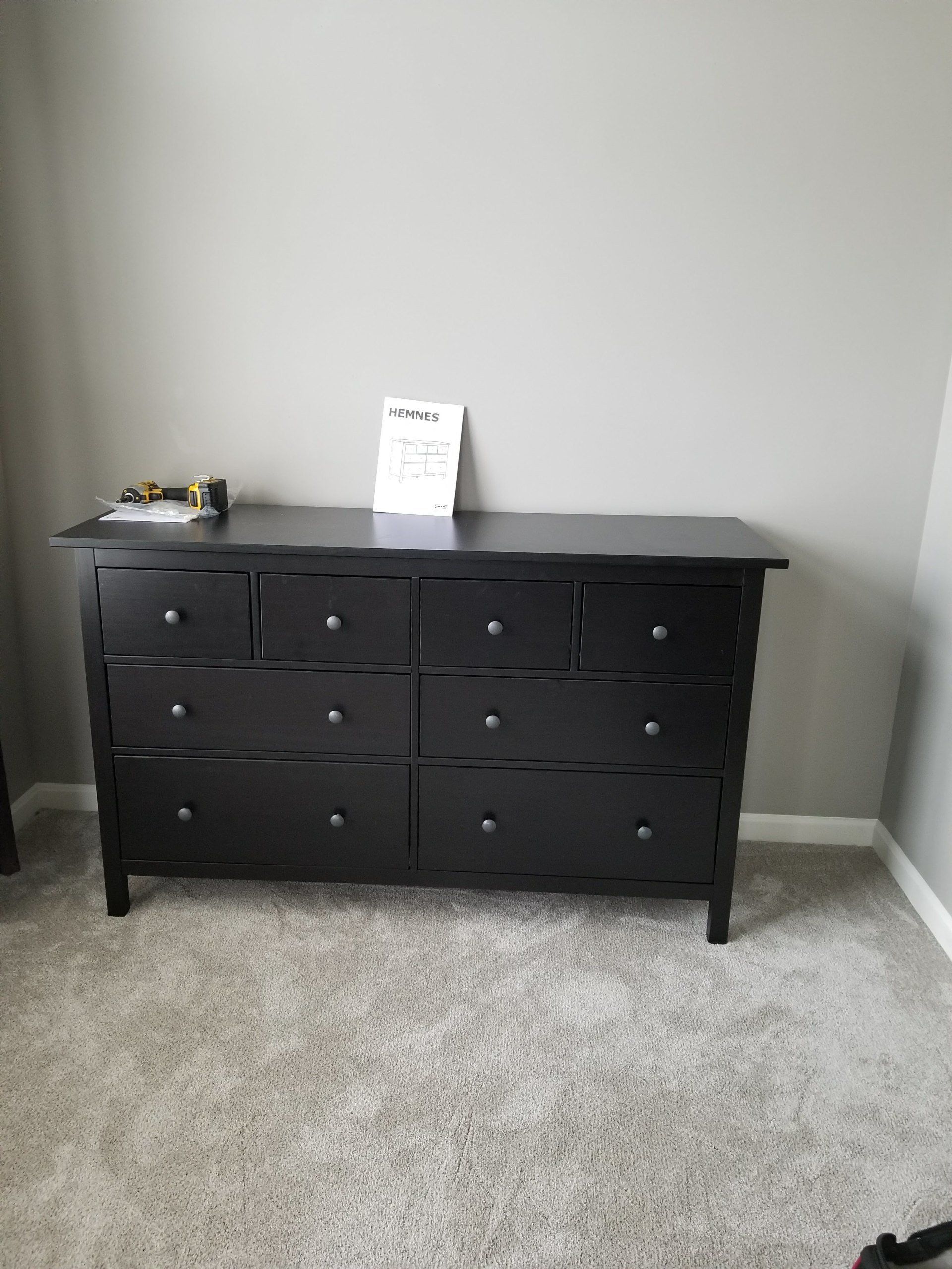 Black dresser with multiple drawers against a gray wall, on gray carpet.