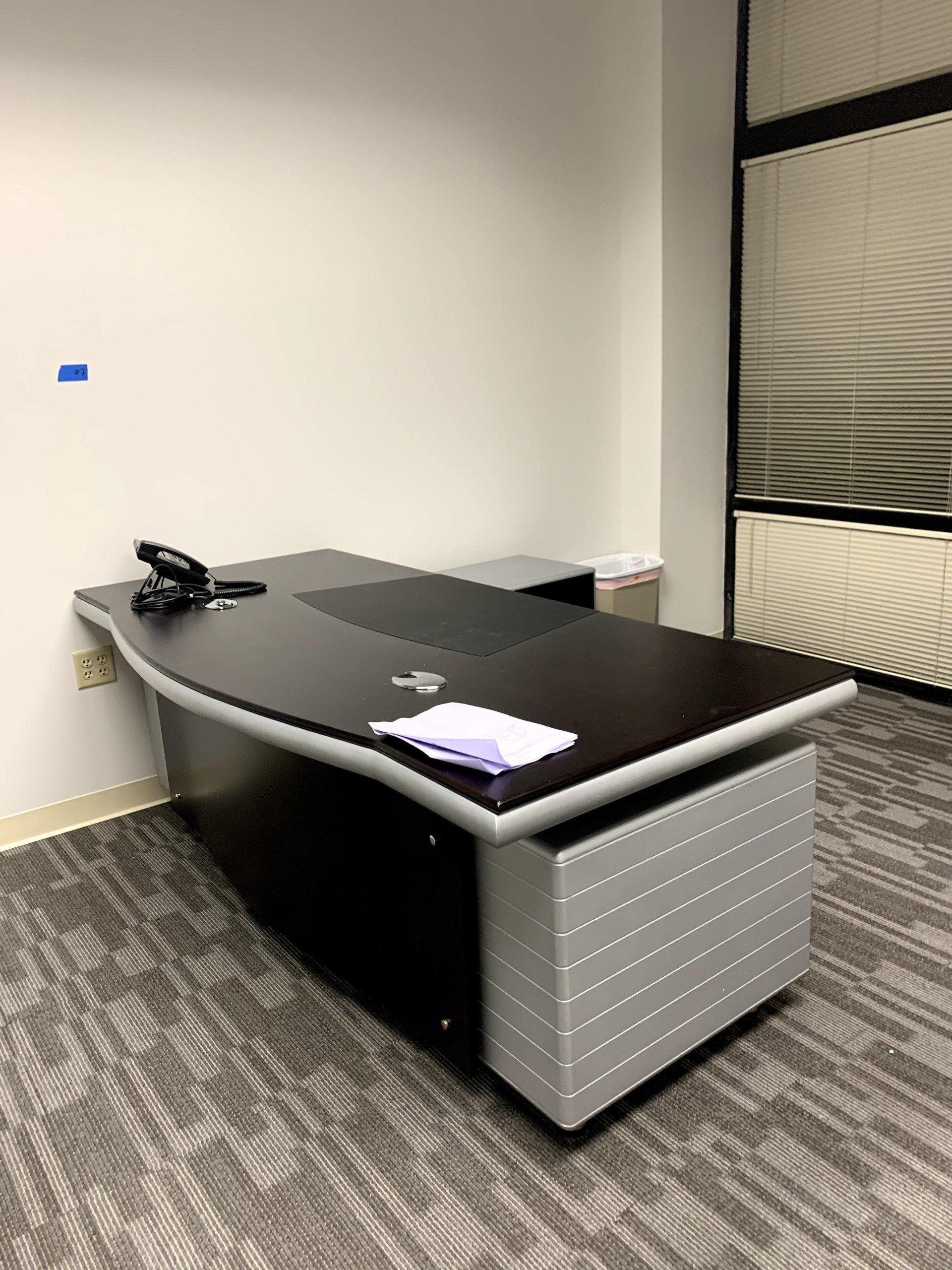 Dark and silver executive desk in an office with white walls and a window with blinds.