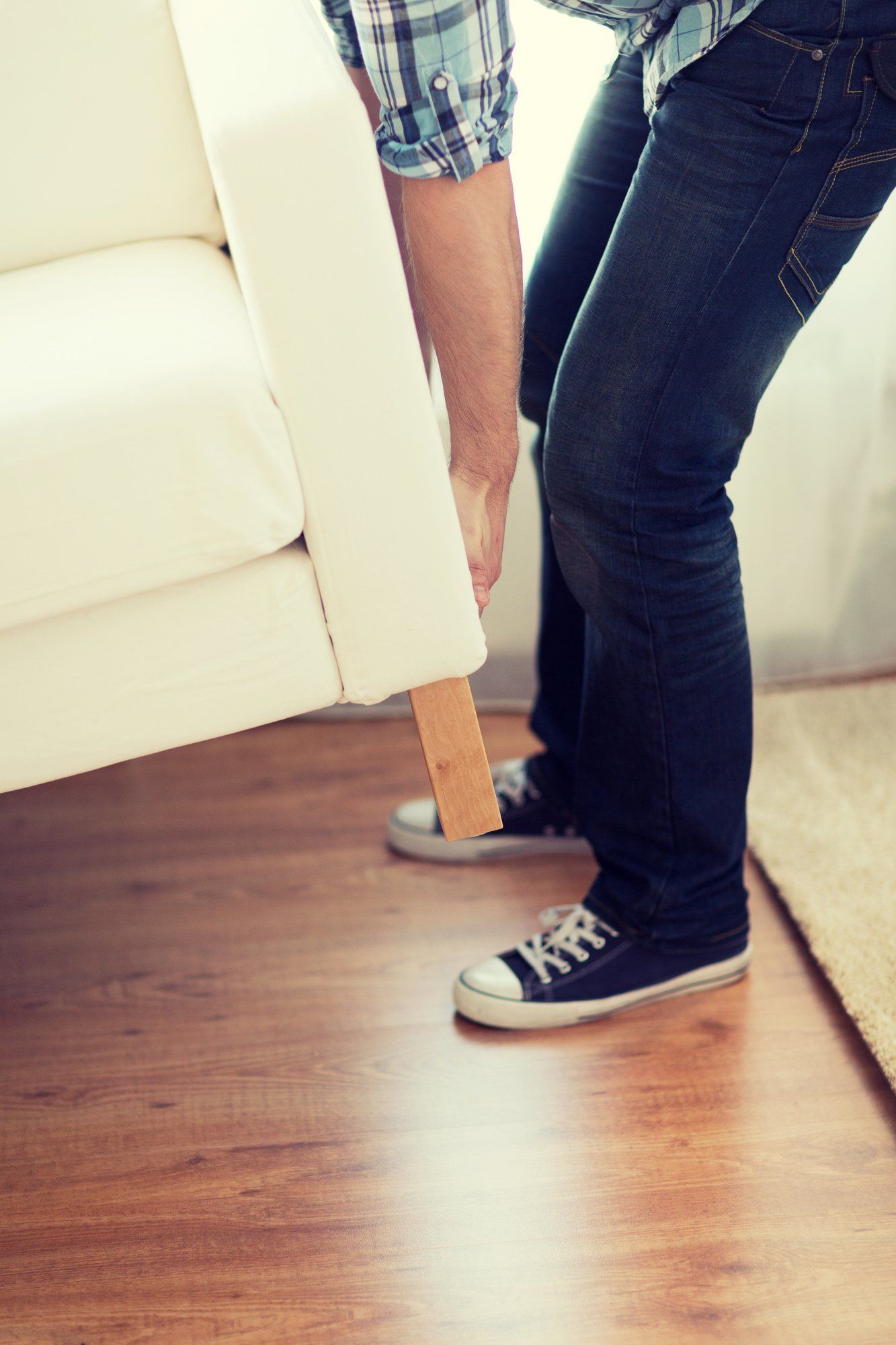 Person in jeans and a plaid shirt lifting a white couch indoors, wooden floor.