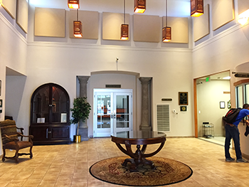 A spacious, brightly lit lobby with tile floors, a circular rug, central table, dark wood cabinet, and a person standing.