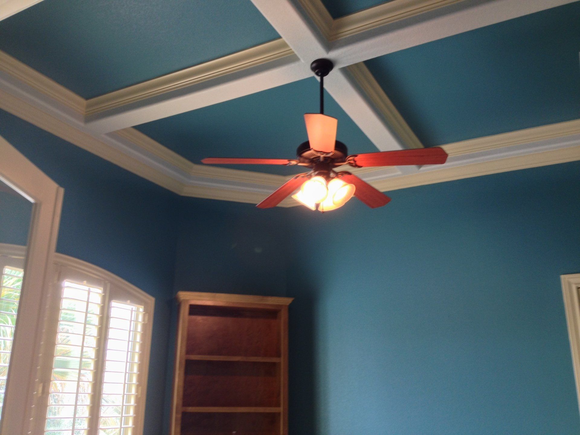A teal-walled room with a coffered ceiling and a wooden ceiling fan featuring a light kit and a corner bookshelf.