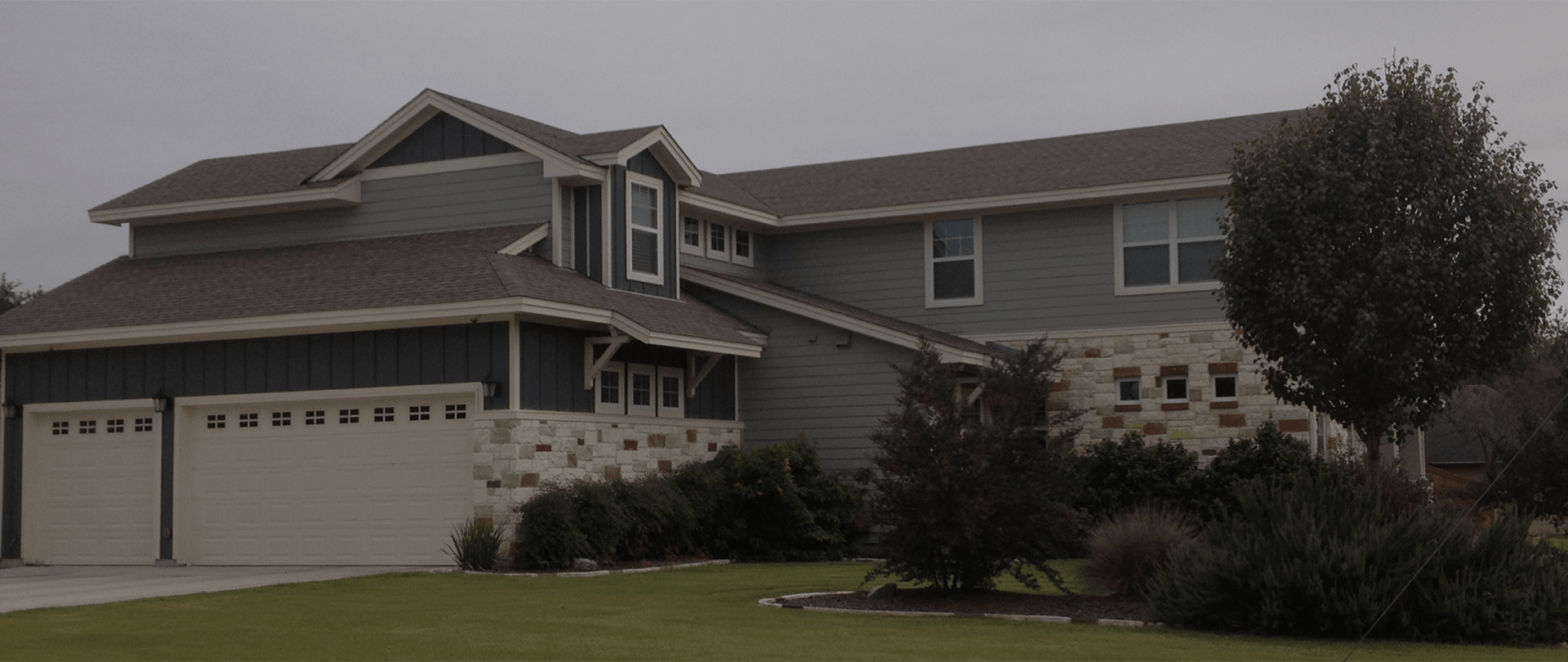 A gray, two-story house with a stone-accented garage and brown shingled roof under an overcast sky.