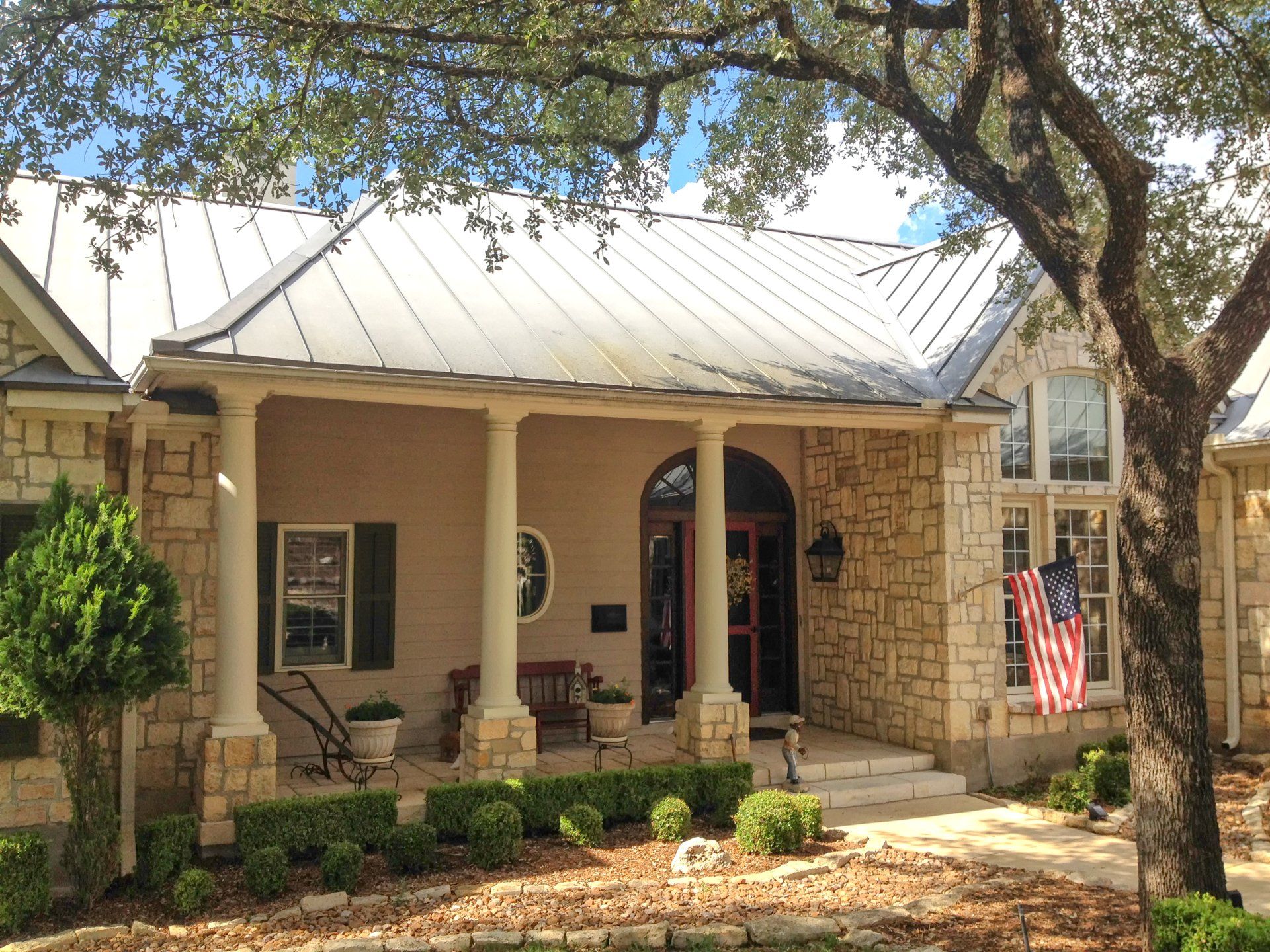 A single-story stone house with a metal roof, a front porch supported by columns, and an American flag near the entryway.