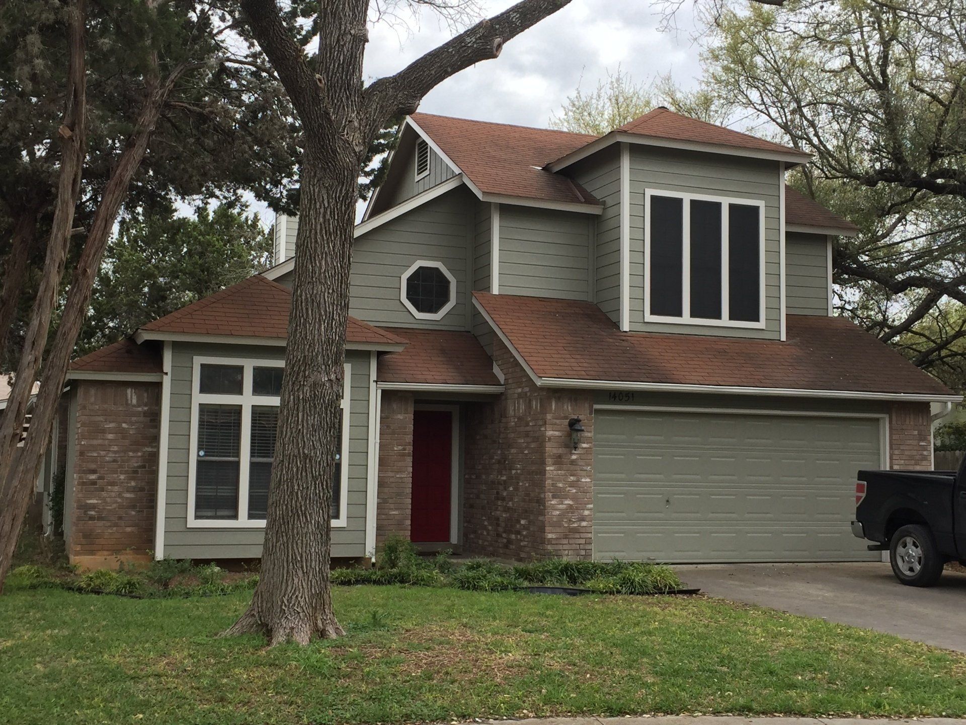A two-story suburban house with olive green siding, brown shingled roof, brick accents, a red door, and a two-car garage.