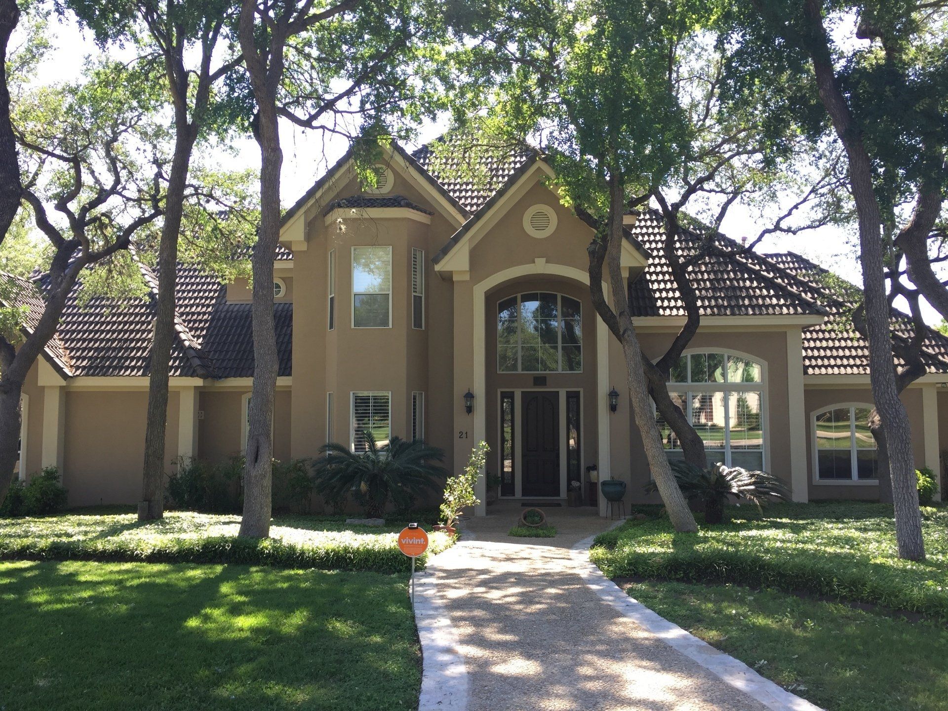 Two-story beige suburban house with a dark brown shingled roof, arched entrance, and gravel path, surrounded by trees.