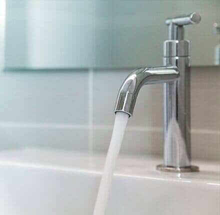 Water Faucet — Willingham Plumbing in VALPARAISO, FL A chrome faucet with running water into a white sink, in front of a tiled wall.