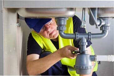 Plumber working under a sink, wearing a reflective vest and using a wrench.